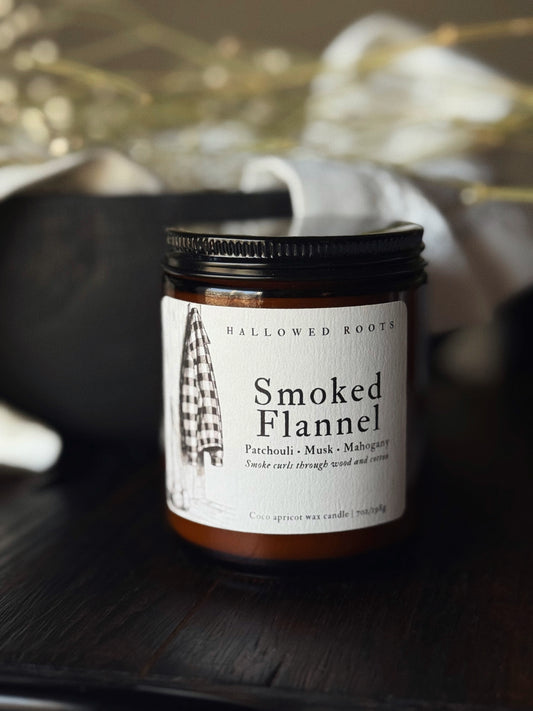 Jar of 'Smoked Flannel' candle by Hallowed Roots on a dark surface with blurred background
