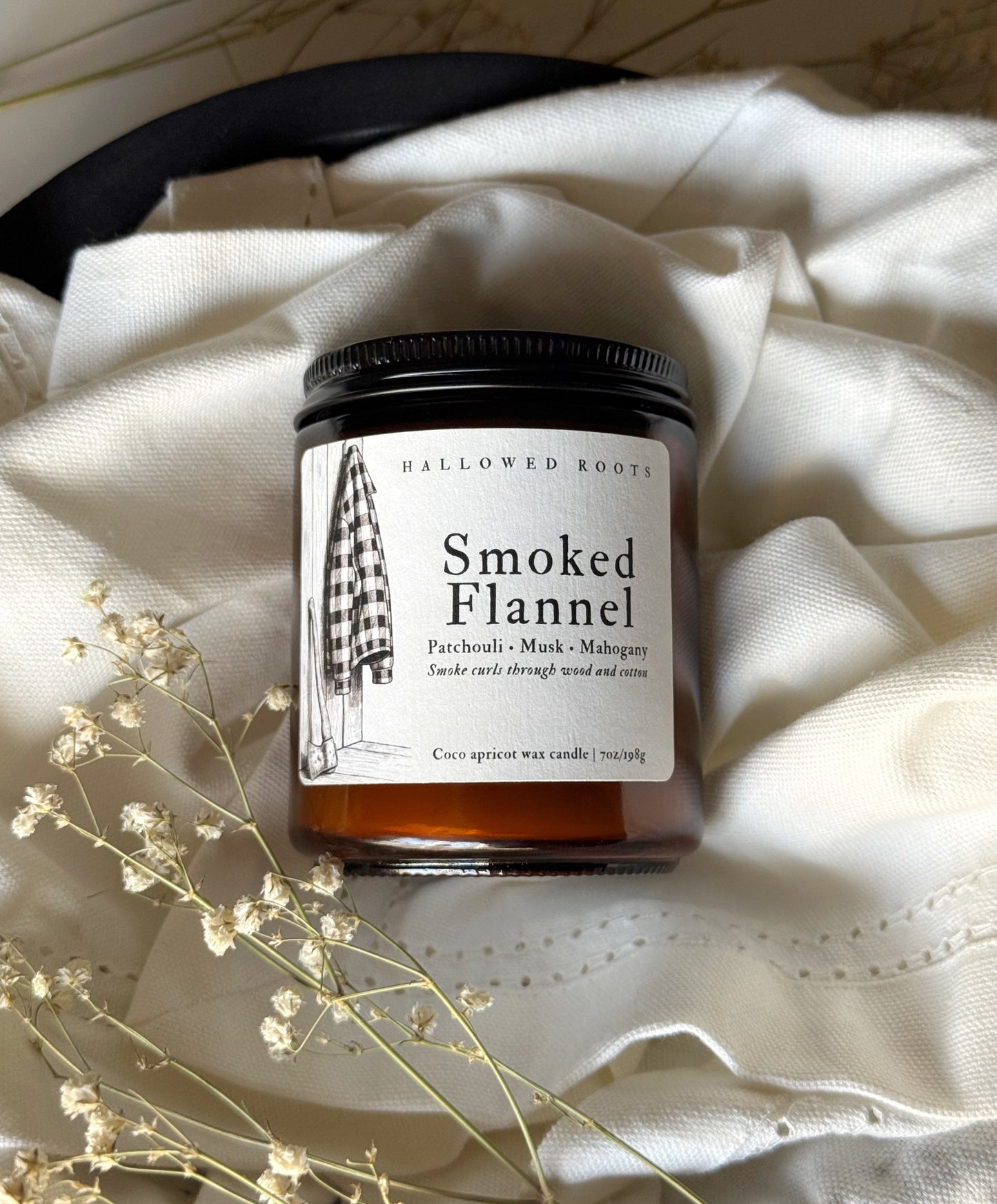 Candle jar labeled 'Smoked Flannel' on a textured surface with dried flowers.