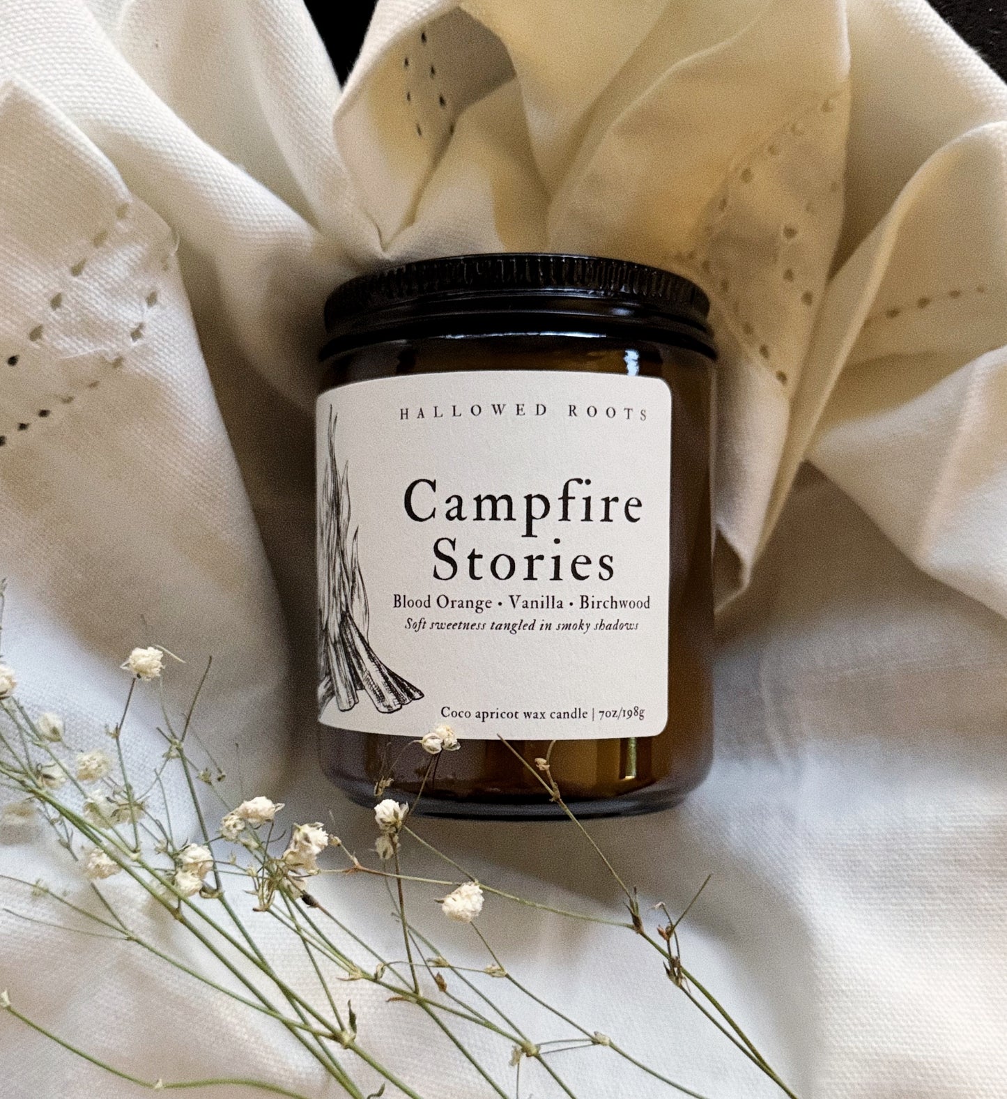 Candle jar labeled 'Campfire Stories' on a textured fabric background with dried flowers.