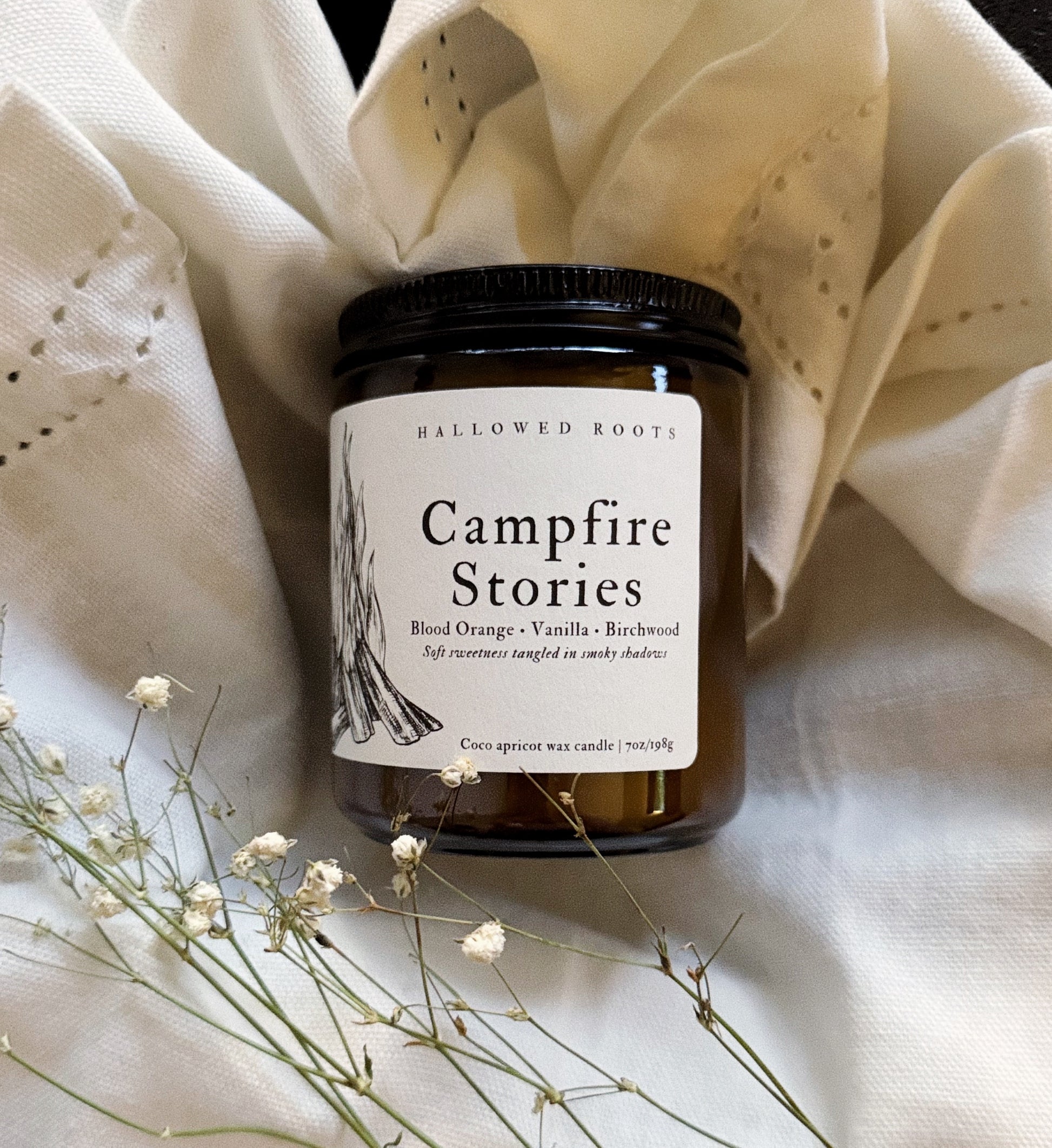 Candle jar labeled 'Campfire Stories' on a textured fabric background with dried flowers.