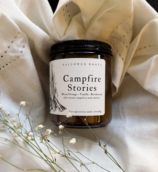Candle jar labeled 'Campfire Stories' on a textured fabric background with dried flowers.
